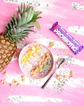 Colorful smoothie bowl with fresh pineapple, probiotics, and sprinkles on a pink background.