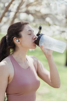 Woman in a tank top drinks water outdoors, rehydrating while wearing earphones.
