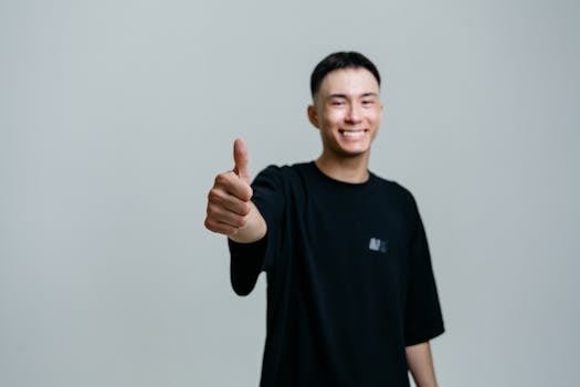 A cheerful young man in a black shirt smiles and gives a thumbs up in a studio setting.