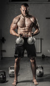 Shirtless man with muscular build lifting kettlebells indoors, showcasing strength and fitness.