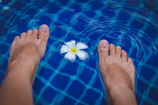 Close-up of feet in a clear blue swimming pool with a floating tropical flower.